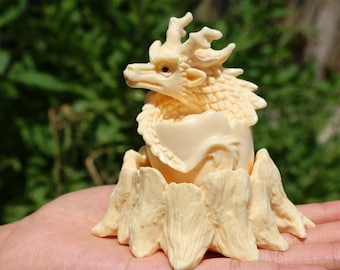 May include: A small, cream-colored dragon sculpture emerging from a cracked egg. The dragon is detailed with scales and has a long, pointed snout. The egg is resting on a base of carved, textured ivory.