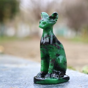 May include: A green and black stone cat figurine. The cat is sitting upright with its ears perked up. The stone has a polished finish, with dark green and black marbling. The figurine is on a gray surface with a blurred background.