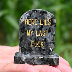 May include: A granite tombstone with the text "HERE LIES MY LAST FUCK" in gold lettering. The stone is dark gray with black and white flecks, and the background is blurred green.