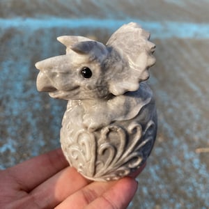 May include: A gray stone sculpture of a baby triceratops emerging from an egg. The dinosaur has a detailed head with horns and a frill. The egg is decorated with swirling patterns. The sculpture is held in a hand.