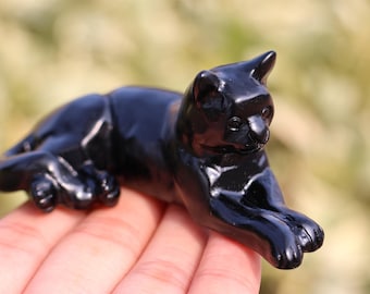 May include: A black obsidian cat figurine, carved in a sleek, realistic style. The cat is lying down with its head turned to the side, and its eyes are closed. The figurine is polished to a high shine, giving it a smooth, glossy finish.