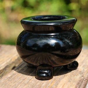 May include: A small, glossy black cauldron-shaped container with three small feet. The container has a rounded body and a wide, open top. It sits on a weathered wooden surface, with a blurred green background.