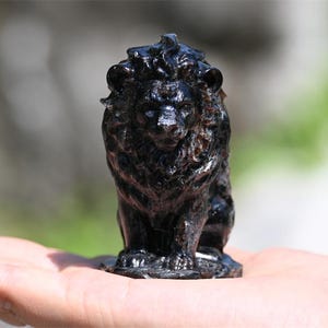 May include: A small, black lion figurine with a detailed mane and a regal pose. The sculpture is held in a hand, showcasing its size and intricate design. The figurine is made of a dark, possibly polished material.