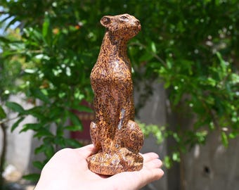 May include: A detailed, brown sculpture of a cheetah standing upright. The cheetah statue has a textured surface with intricate patterns. The sculpture is held in a hand against a backdrop of green foliage.