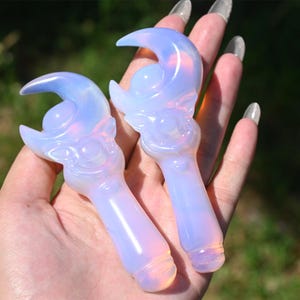 Pode incluir: Duas varinhas de cristal opalescente, cada uma com um design de lua crescente. As varinhas têm uma base arredondada e são seguradas em uma mão contra um fundo verde desfocado. As varinhas têm uma aparência suave e brilhante, com cerca de 10 cm de comprimento.