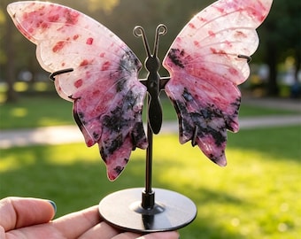 Puede incluir: Una talla con forma de mariposa hecha de piedra rosa y negra, montada sobre un soporte fino de metal negro con base circular. La piedra presenta zonas de color rosa translúcido con inclusiones negras oscuras y moteadas en las alas.