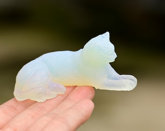 May include: A translucent, milky white cat figurine carved from opalite. The sculpture depicts a cat in a resting, lying-down position with its front paws extended forward and its head turned slightly to the side, showing smooth, curved lines.