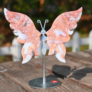 Puede incluir: Una escultura de mariposa de metal con un acabado marmoleado blanco y naranja. La mariposa está de pie sobre un soporte de metal plateado con una base redonda.
