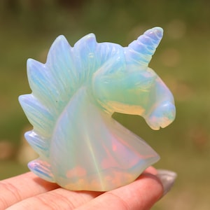 May include: A carved unicorn figurine in opalescent colors of white, blue, and yellow. The sculpture is held in a hand and has a detailed mane and horn. The background is blurred, suggesting an outdoor setting.