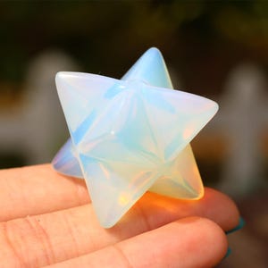 Puede incluir: Un cristal blanco iridiscente en forma de estrella, también conocido como Merkaba, se sostiene en una mano. El cristal está hecho de ópalo y tiene una apariencia translúcida.