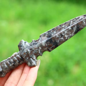 Puede incluir: Una daga de piedra tallada con un patrón moteado en tonos grises, marrones y blancos. El mango tiene un diseño detallado y texturizado. La hoja es puntiaguda y la forma general es simétrica. El fondo es verde borroso.