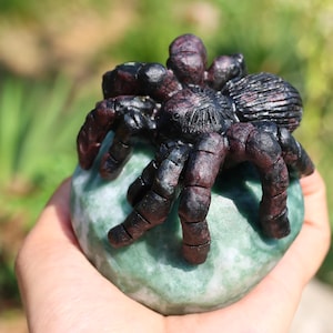 May include: A detailed sculpture of a spider with a dark body and legs, perched on a light green stone. The spider's body is a deep black, with reddish-brown legs. The stone has a mottled appearance.