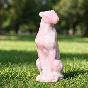 May include: A carved stone figurine of a seated panther, featuring a smooth, polished surface with a pale pink and white marbled appearance. The feline sculpture is depicted in an upright, alert posture with its head turned to the right.