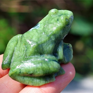Pode incluir: Uma estatueta de rã verde esculpida. A rã é de uma cor verde vibrante com veios verdes mais escuros. A rã está sentada, com as pernas dobradas. A estatueta é segurada na mão de uma pessoa.