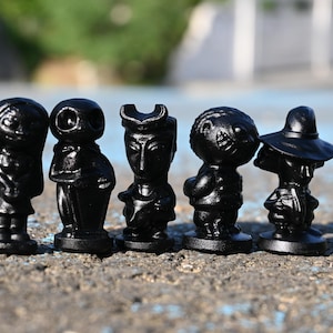 May include: Five small, black figurines of different characters are arranged in a line. The figurines include a figure with a hat, a figure with a hat and a dress, and other distinct shapes.