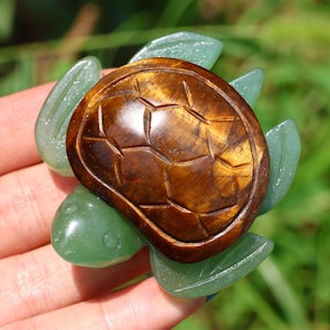 May include: A handcrafted turtle figurine with a brown, patterned shell and green, leaf-shaped flippers. The turtle is made of polished stone and is held in a hand against a blurred green background.
