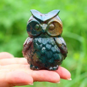 May include: A small, carved owl figurine in shades of green, brown, and burgundy. The owl has large, round eyes and a textured surface. The figurine is held in a hand against a blurred green background.