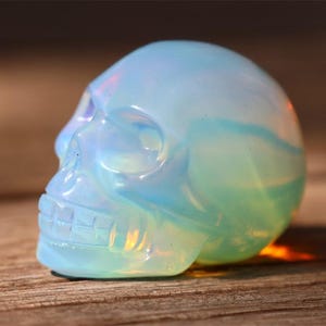 May include: A translucent, opalescent skull figurine with a light blue and green color gradient. The skull is carved with detailed features, including eye sockets, a nose cavity, and teeth. The figurine is displayed on a wooden surface.