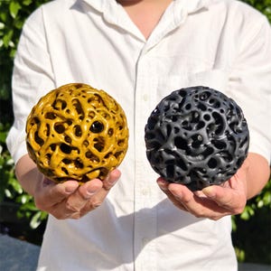 May include: Two spherical decorative objects. One is a golden yellow color, and the other is dark gray. Both have a porous, organic design with numerous holes throughout their surface. The objects are held in front of a green leafy background.
