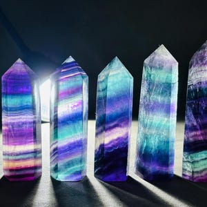May include: Five polished fluorite crystal towers. Each tower is a different shade of purple, blue, and green, with a striped pattern. The crystals are standing upright on a dark surface, with a light source behind them.