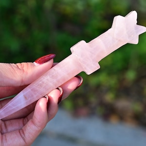 Può includere: Una bacchetta di quarzo rosa rosa chiaro con una parte superiore a forma di stella. La bacchetta è tenuta in mano su uno sfondo verde sfocato. La bacchetta è lunga circa 15 cm.