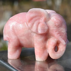 May include: A small pink carved elephant figurine. The elephant is standing on a silver surface.