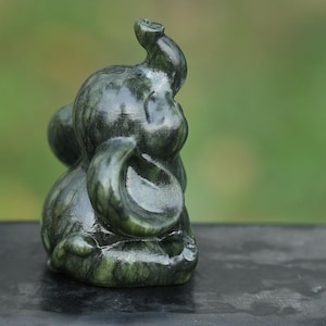May include: A small, dark green elephant figurine with a curled trunk and large ears. The sculpture has a smooth, polished finish and is set against a blurred green background. The elephant is a symbol of good luck and fortune.