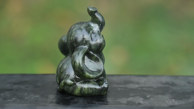 May include: A small, dark green elephant figurine with a curled trunk and large ears. The sculpture has a smooth, polished finish and is set against a blurred green background. The elephant is a symbol of good luck and fortune.