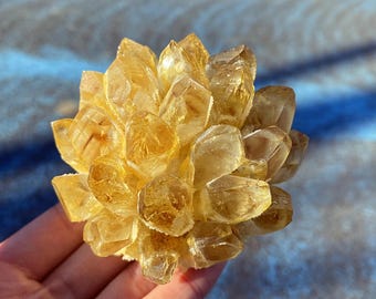 May include: A cluster of golden-yellow crystals, resembling a flower or starburst. The crystals have a translucent quality, catching the light and displaying various facets. The overall shape is rounded and compact, with a natural, organic aesthetic.