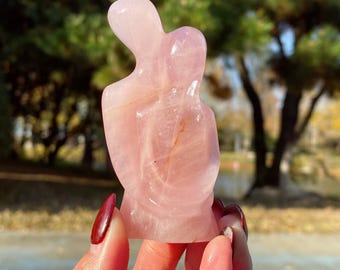 May include: A pink rose quartz sculpture depicting two figures embracing. The polished stone has a smooth surface and a soft, translucent appearance. The sculpture is held in a hand with red nail polish, with a blurred outdoor background.