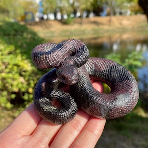 Könnte beinhalten: Eine detaillierte Skulptur einer sich windenden Schlange in Lila- und Schwarztönen. Die Schuppen der Schlange sind aufwendig geschnitzt, und ihr Kopf ist erhoben. Die Skulptur wird in einer Hand gehalten, mit einem verschwommenen Hintergrund im Freien.