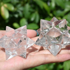 May include: Two clear quartz Merkaba stars, each with multiple triangular facets, are held in a hand. The stars are transparent, allowing light to pass through. The background is blurred green foliage.