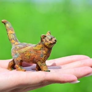 Puede incluir: Una pequeña figura de gato pintada a mano con una superficie texturizada y un patrón multicolor en tonos naranja, amarillo, marrón y verde. El gato se muestra en posición de pie, con la cola levantada y las orejas apuntando hacia adelante.