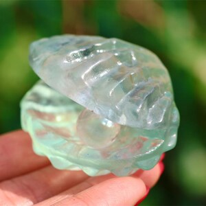 May include: A translucent, carved gemstone clam shell with a pearl inside. The shell is a pale green color with a textured surface. The clam shell is held in a person's hand against a blurred green background.
