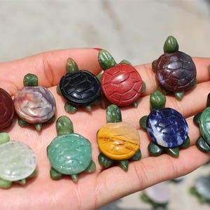 May include: A collection of small, carved stone turtle figurines in various colors, including red, green, black, and yellow. The turtles are held in a hand, showcasing their detailed shell patterns and green heads and legs. These decorative figurines are ideal for collectors.