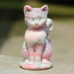May include: A carved cat figurine in shades of pink and white. The cat is sitting upright with its tail curled. The stone sculpture has a smooth, polished finish and is set on a round base. The cat's features are detailed, with visible ears and a gentle expression.