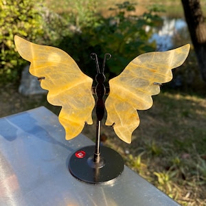 May include: A decorative butterfly sculpture with translucent yellow wings and a dark metal body. The wings have a textured appearance and are mounted on a black metal stand. The sculpture is displayed on a silver surface.