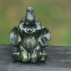 May include: A dark green elephant figurine with a trunk raised upwards and large ears. The sculpture is made of a stone-like material and sits on a dark surface. The background is blurred green.