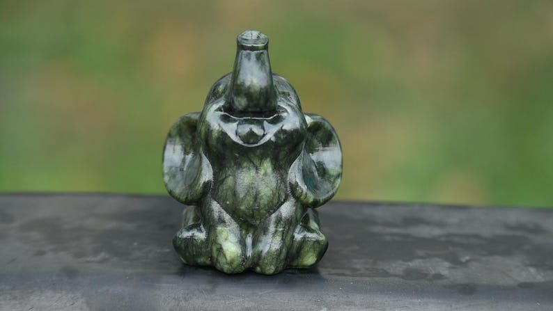 May include: A dark green elephant figurine with a trunk raised upwards and large ears. The sculpture is made of a stone-like material and sits on a dark surface. The background is blurred green.