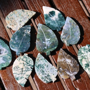 May include: A collection of leaf-shaped stones in shades of green, white, and brown, arranged on a wooden surface. The stones vary in pattern and texture, with some displaying a marbled appearance. The natural stone leaves are a decorative item.