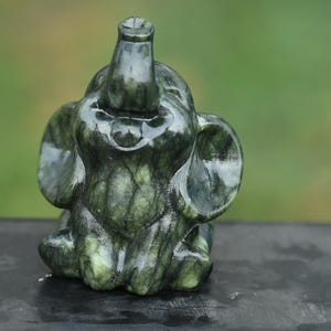 May include: A small, dark green elephant figurine with a trunk raised upwards and large ears. The stone sculpture has a polished, glossy finish and sits on a dark surface. The background is a blurred green.