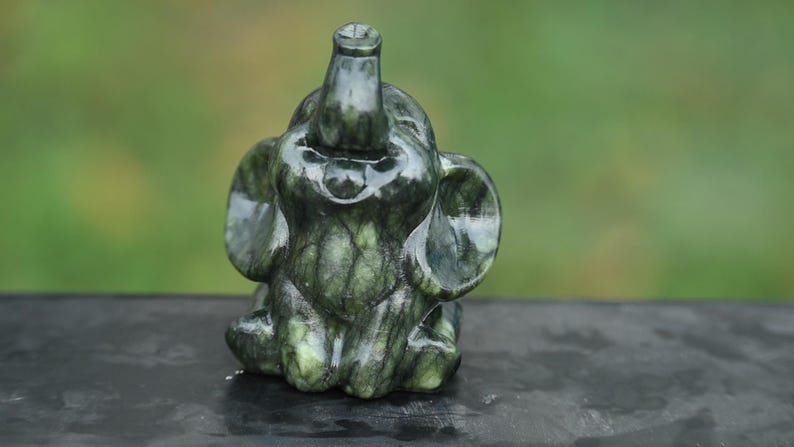 May include: A small, dark green elephant figurine with a trunk raised upwards and large ears. The stone sculpture has a polished, glossy finish and sits on a dark surface. The background is a blurred green.