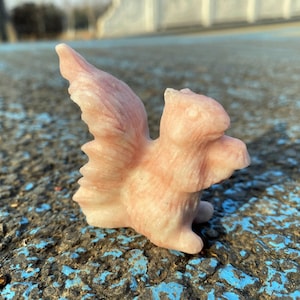 Puede incluir: Una figura de ardilla rosa tallada con los brazos extendidos y una cola tupida. La ardilla está hecha de una piedra rosa claro y tiene una textura de pelaje detallada. La figura está sobre una superficie azul y negra.
