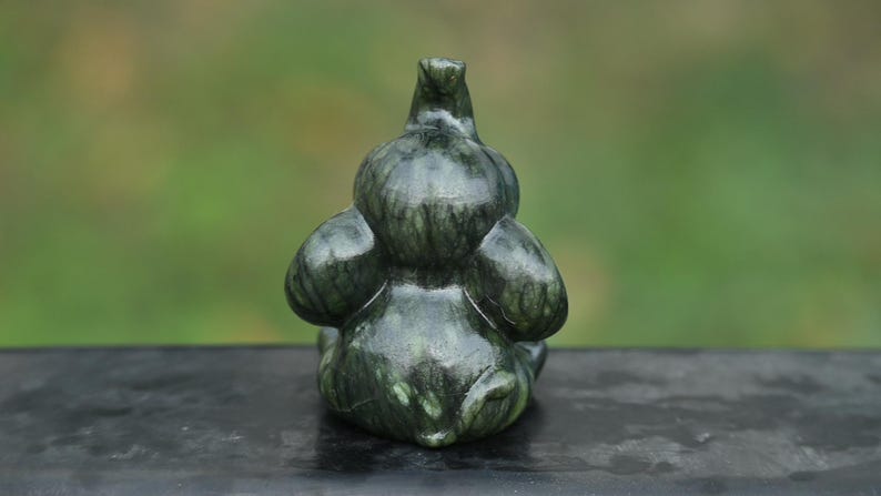May include: A dark green, carved stone sculpture of a duck or bird. The sculpture has a smooth, polished appearance with visible veining. The bird is depicted in a seated position, with its wings folded and head upright. The background is blurred.