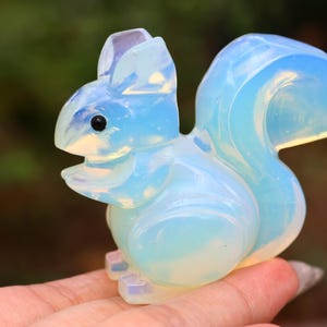 Puede incluir: Una figura de ardilla opalina tallada en azul claro y blanco. La escultura tiene un acabado liso y pulido y ojos negros. La ardilla está en posición sentada con la cola curvada hacia arriba. Un artículo decorativo.