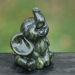 May include: A small, dark green elephant figurine with its trunk raised. The sculpture has a smooth, polished surface and is sitting down. The elephant's ears are large and rounded, and the overall design is detailed.
