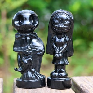 Op de afbeelding: Twee zwarte beeldjes, een mannelijk en een vrouwelijk, staan naast elkaar. De mannelijke figuur draagt een jas en heeft een schedelachtig gezicht. De vrouwelijke figuur heeft lang haar en een brede grijns. Beide staan op ronde voetstukken.