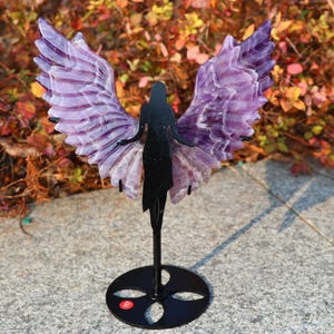 May include: A purple amethyst crystal angel wing sculpture on a black metal stand. The angel is depicted in silhouette with wings made of amethyst.