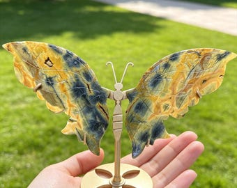 May include: A decorative butterfly sculpture featuring wings carved from yellow and blue stone. The butterfly is mounted on a gold-colored metal stand with a circular base that has a four-petal cutout design, held upright by a central vertical rod.