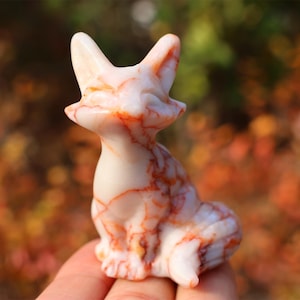 Puede incluir: Una figurita de zorro blanco y rojo tallada en una piedra de mármol rojo y blanco. La figurita está sentada en una mano.